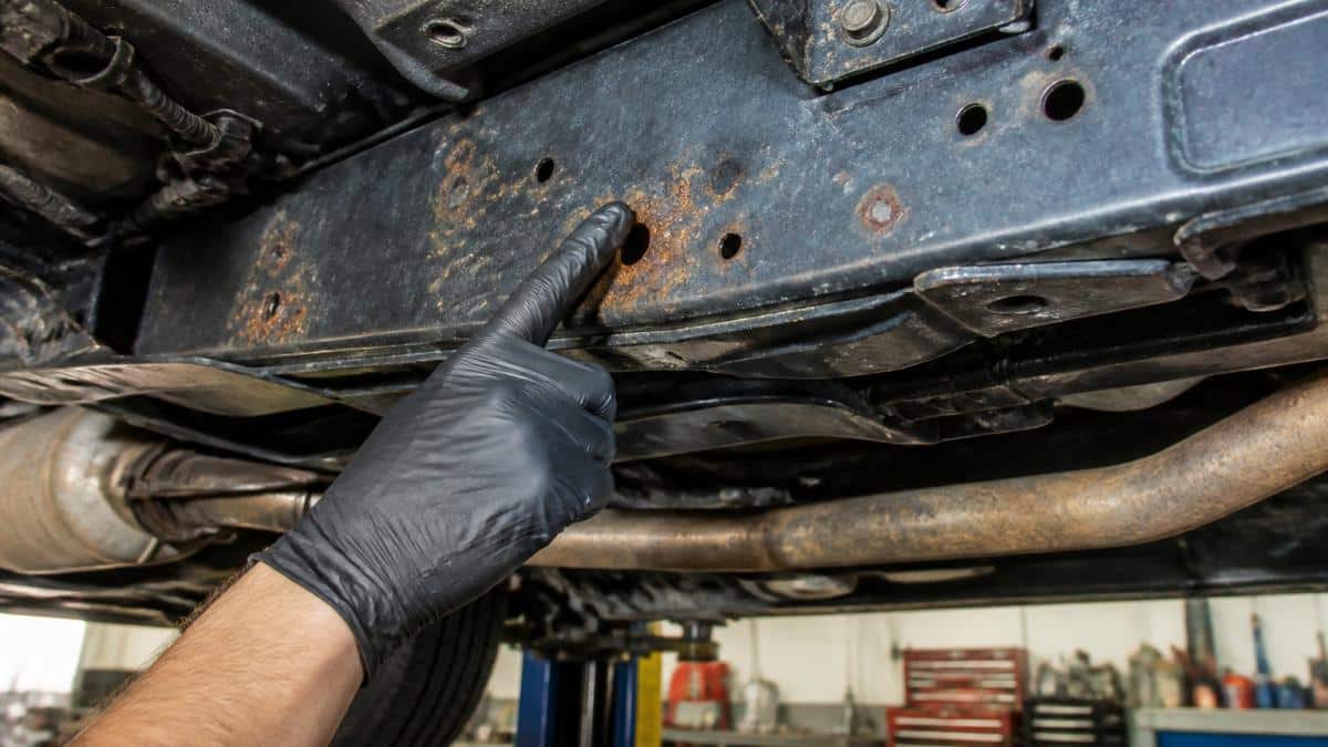 Close-up of a vehicle's undercarriage showing early rust spots on a metal frame, with a technician's gloved hand pointing at the affected area in a well-lit garage